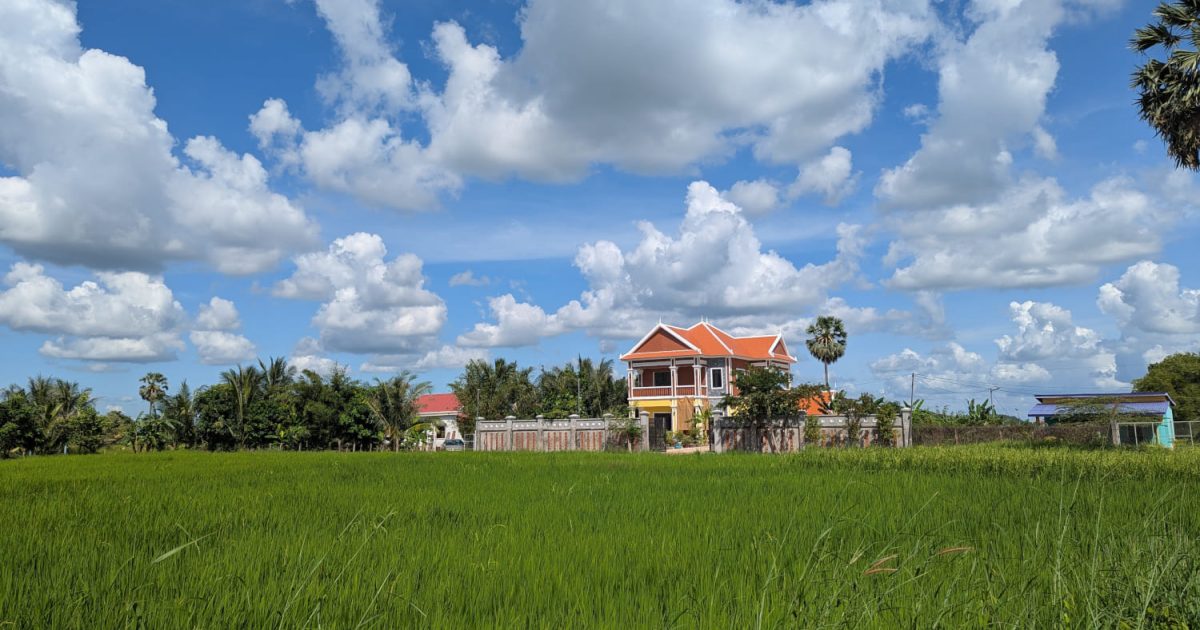 Haus und Reisfeld in Kep, Kambodscha Traditionelles kambodschanisches Haus mit orangefarbenem Dach inmitten grüner Reisfelder unter blauem Himmel - sicheres ländliches Kambodscha.